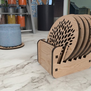 Puede incluir: Un juego de seis posavasos de madera con un diseño de hojas superpuestas cortado con láser. Los posavasos están apilados en un soporte de madera.