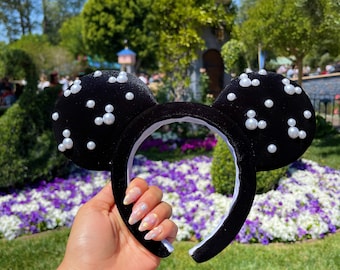 Pode incluir: Orelhas de Minnie Mouse de veludo preto com enfeites de pérolas brancas. As orelhas são seguradas em uma mão com esmalte de unha colorido.