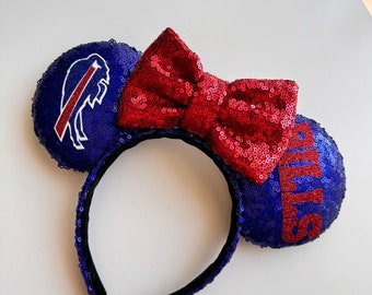 Pode incluir: Orelhas de Minnie Mouse de lantejoulas azuis e vermelhas com um laço vermelho e um logotipo do Buffalo Bills em uma orelha. A outra orelha tem a palavra "Bills" em vermelho.