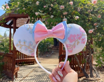 Pode incluir: Faixa de cabeça de orelhas de rato brancas com um laço rosa e azul. As orelhas são bordadas com desenhos florais em rosa e azul.