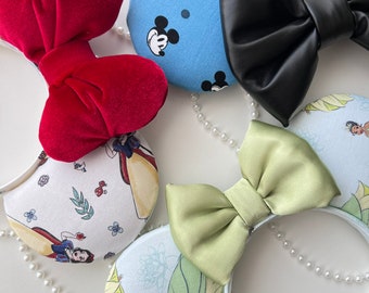 Pode incluir: Quatro pares de orelhas de Minnie Mouse com tema de personagens da Disney com laços e padrões de tecido de diferentes cores. As orelhas são brancas com uma variedade de desenhos de personagens da Disney, incluindo Branca de Neve, Tiana, Mickey Mouse e um laço de veludo vermelho.