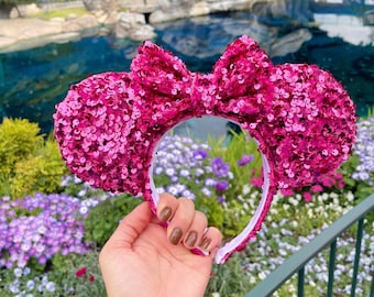 Pode incluir: Faixa de cabeça de orelhas de Minnie Mouse com lantejoulas rosa e um grande laço. A faixa de cabeça é segurada em uma mão com esmalte de unha rosa.