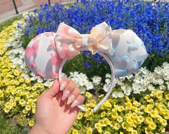 Pode incluir: Faixa de cabeça branca com duas orelhas de tecido decoradas com borboletas rosa pastel, azul e pêssego. A faixa de cabeça é segurada na frente de um leito de flores coloridas.