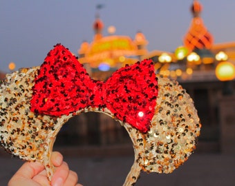 Pode incluir: Orelhas de Minnie Mouse de lantejoulas douradas com um laço de lantejoulas vermelhas. As orelhas estão em foco, enquanto o fundo está desfocado.