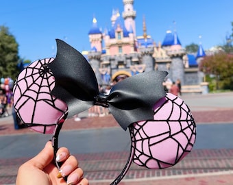 Pode incluir: Orelhas de Minnie Mouse rosa e pretas com um design de teia de aranha e um laço preto. As orelhas estão na frente de um castelo rosa e azul.