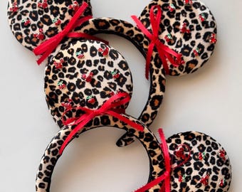Pode incluir: Um par de orelhas de rato de estampa de leopardo com laços vermelhos e strass em forma de cereja. A faixa de cabeça é preta.