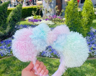 Pode incluir: Um par de orelhas de Minnie Mouse felpudas em azul pastel e rosa com uma faixa de cabeça branca. As orelhas são em um padrão tie-dye.