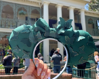 Pode incluir: Faixa de cabeça verde com um grande laço e enfeites de borboleta. A faixa de cabeça é segurada na frente de um edifício com colunas brancas.