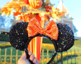 Pode incluir: Faixa de cabeça de orelhas de Minnie Mouse com lantejoulas pretas e laranja com um grande laço laranja. A faixa de cabeça é segurada na frente de um cenário festivo de Halloween.