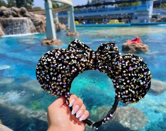 May include: Black and gold sequined Minnie Mouse ears headband with a black velvet bow. The headband is held in front of a blue pool with a waterfall in the background.