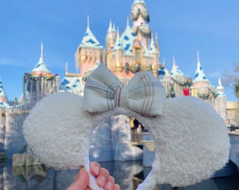 Pode incluir: Orelhas de Minnie Mouse brancas e felpudas com um laço xadrez em tons de bege, marrom e branco. As orelhas estão na frente de um castelo azul e rosa com detalhes brancos.