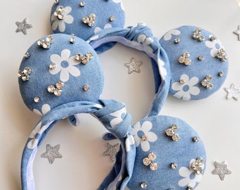 Pode incluir: Faixa de cabeça de denim azul com estampa de margaritas brancas e strass prateados. A faixa de cabeça é amarrada em um laço na parte superior.