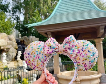 Pode incluir: Orelhas de Minnie Mouse cor-de-rosa com um grande laço coberto de lantejoulas coloridas. As orelhas estão na frente de uma fonte de pedra com um gazebo no fundo.