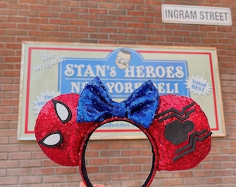 Pode incluir: Orelhas vermelhas de Spider-Man com lantejoulas, detalhes de olhos brancos e laço azul com lantejoulas. A faixa de cabeça é preta. O fundo inclui uma placa que diz "Stan's Heroes New York Deli" e uma parede de tijolos.