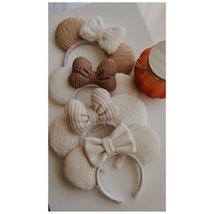 May include: A set of four pairs of mouse ears with different fabric textures and bow styles. The ears are in shades of beige, brown, and white.