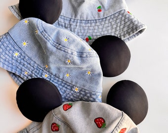 Pode incluir: Três chapéus de pescador de brim com orelhas de Mickey Mouse pretas. Os chapéus têm diferentes desenhos bordados: morangos, margaridas e morangos.