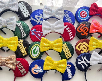 Pode incluir: Uma coleção de 15 pares de orelhas de Minnie Mouse com tema esportivo, de lantejoulas e cores vibrantes. Cada par apresenta um logotipo de equipe diferente, incluindo os Raiders, os Eagles, os Steelers, os Packers, os Dodgers, os Angels, os Cubs, os Giants, os Yankees, os 49ers, os Rams, e mais.