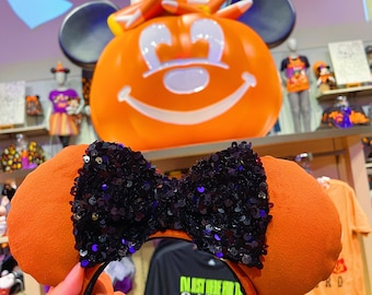 Pode incluir: Orelhas de Minnie Mouse laranja e pretas com um laço preto de lantejoulas. As orelhas têm o formato das orelhas do Mickey Mouse e têm uma faixa de cabeça.