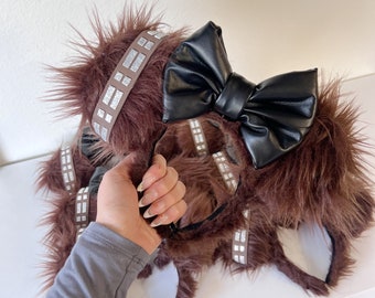 Pode incluir: Faixa de cabelo marrom peluda inspirada em Chewbacca com um laço de couro falso preto. A faixa de cabelo tem detalhes de glitter prata.