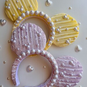 May include: A pair of purple and yellow mouse ears with pearl embellishments. The ears are made of a soft, textured fabric and have a headband.