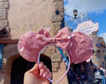 Pode incluir: Orelhas de Minnie Mouse cor-de-rosa com um laço e detalhes em tecido em forma de borboleta. As orelhas são seguradas na frente de um prédio rosa com uma placa que diz "Main Street Boutique".
