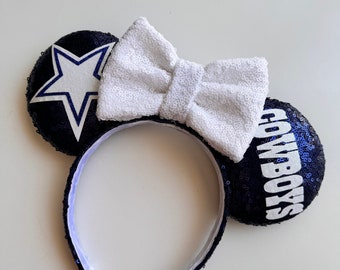 Pode incluir: Orelhas de rato com lantejoulas azul marinho com um laço branco com lantejoulas. Uma orelha apresenta uma estrela branca sobre um fundo azul marinho, a outra a palavra "Cowboys" em branco. A tiara é azul marinho.