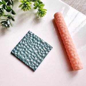 Può includere: Un quadrato testurizzato color turchese e un rullo testurizzato rosa, entrambi con un motivo a pelle di coccodrilo, utilizzati per creare disegni in rilievo nell'argilla.