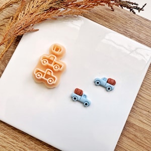 May include: A light peach-colored cookie cutter in the shape of a truck carrying a pumpkin. Two small blue trucks with pumpkins in the back are also present. The items are on a white surface with a wooden background.