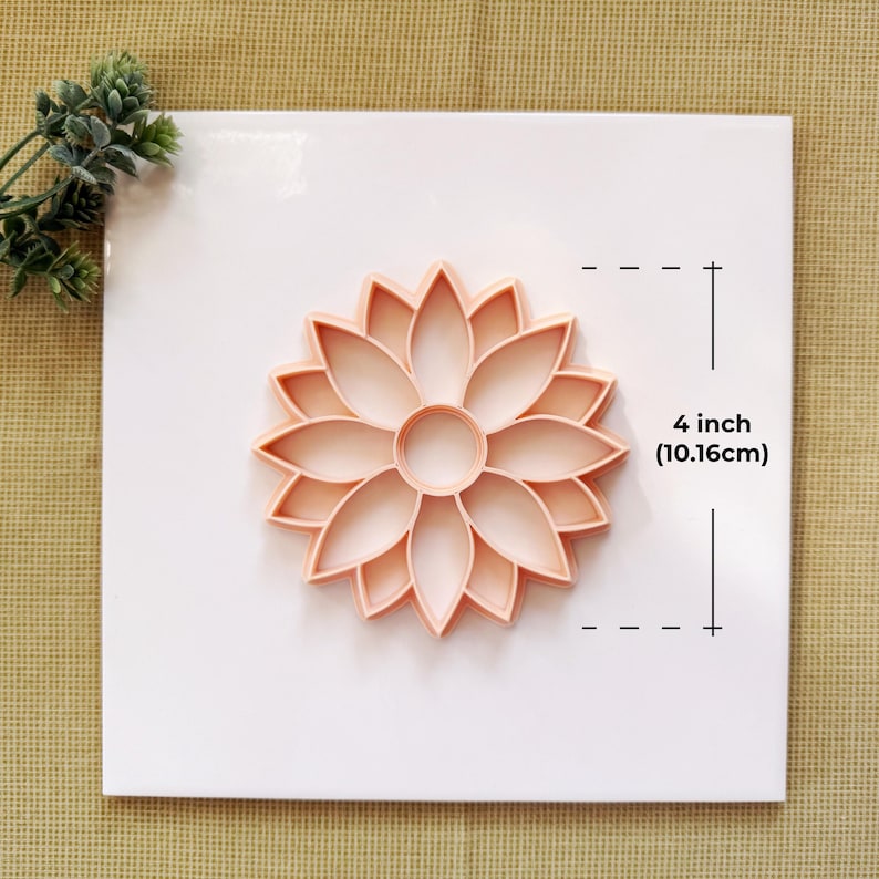 May include: A light peach-colored flower-shaped cookie cutter, measuring 4 inches (10.16cm) in diameter, is displayed on a white surface. The cutter features a detailed petal design, perfect for baking. A sprig of greenery is visible in the upper left corner.