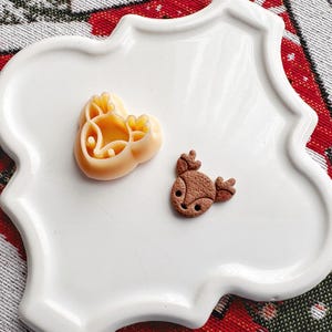 Könnte beinhalten: Zwei rentierförmige Ausstecher auf einem weißen Teller. Einer ist aus braunem Teig, der andere aus gelbem Teig.