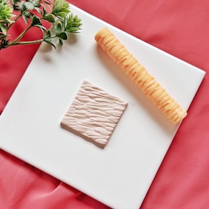 May include: A square, textured, light brown stamp and a long, ridged, orange-yellow roller are displayed on a white square surface. The items are arranged diagonally, with a sprig of green leaves in the upper left corner.