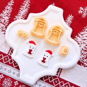 May include: A white, decorative plate holds Christmas-themed cookie cutters and decorated cookies. The cutters are in the shape of a bell and a gift. The cookies are white ghosts wearing Santa hats and holding red gifts. The plate sits on a red and white patterned surface.
