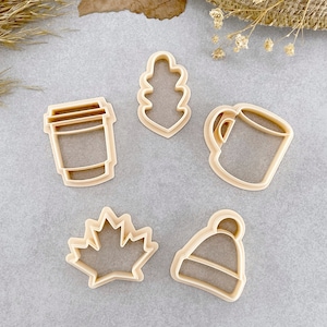 May include: Five cookie cutters made of beige plastic. The cutters are shaped like a coffee cup, a leaf, a maple leaf, a toque, and a to-go coffee cup.