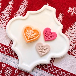 Op de afbeelding: Drie hartvormige koekjessnijders in oranje, roze en perzik, elk met een bloemenontwerp. Ze liggen op een witte, decoratieve, geschulpte plaat tegen een rood-witte achtergrond.