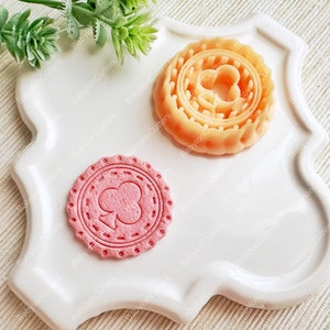 Könnte beinhalten: Zwei Ausstecher mit einem Kleeblatt-Design. Einer ist hellorange, der andere rosa. Sie liegen auf einem weißen Teller mit gewelltem Rand. Der Text "Babylon Cutters" ist im Hintergrund sichtbar.