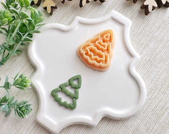May include: Two Christmas tree-shaped cookie cutters on a white ceramic plate. One cutter is orange and the other is green.