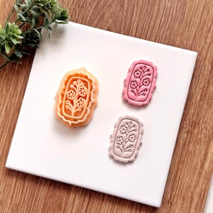 Może przedstawiać: Trzy wykrawacze do ciastek w kształcie kwiatów w odcieniach pomarańczu, różu i fioletu. Każdy wykrawacz ma prostokątną ramkę z falistą krawędzią i kwiatowym wzorem. Wykrawacze są ułożone na białej powierzchni z drewnianym tłem.