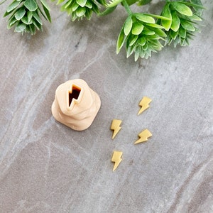 May include: A set of five small, yellow lightning bolt shaped charms. The charms are on a light gray surface. There is a small, beige, 3D printed object with a lightning bolt cutout in the center.