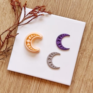 May include: Three crescent moon-shaped objects featuring lunar phase patterns. One is a light peach colour with a smooth finish, one is a deep purple with a textured surface, and one is a light grey with a textured surface, all arranged on a white tile.