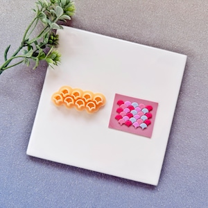Könnte beinhalten: Ein orangefarbener, muschelförmiger Ausstecher und ein rosa Quadrat mit einem Mosaik aus rosa, roten und blauen muschelförmigen Fliesen. Die Artikel befinden sich auf einer weißen quadratischen Fliese, mit einem Zweig grüner Blätter im Hintergrund.