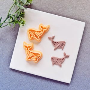 May include: Four whale-shaped cookie cutters. Two are orange with floral details, and two are mauve with floral designs. The cutters are on a white square plate, with a sprig of greenery in the upper left corner.