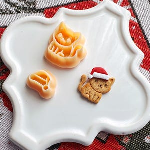 May include: A white ceramic plate with a scalloped edge features three cookie cutters shaped like cats. One cutter is a simple cat shape, another is a cat wearing a Santa hat, and the third is a cat with a heart on its chest.