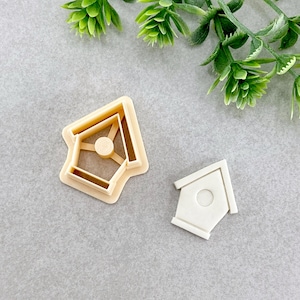 May include: Two plastic cookie cutters in the shape of a birdhouse. The larger cutter is beige and the smaller cutter is white.