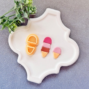 Può includere: Un vassoio decorativo bianco contiene tre ciondoli a forma di cono gelato nei toni del rosa, marrone e arancione. Il vassoio contiene anche due ciondoli arancioni a forma di vongola. Un rametto di fogliame verde si trova nell'angolo in alto a sinistra.