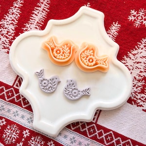 May include: A white, decorative, quatrefoil-shaped plate holds four bird-shaped molds. Two are orange with floral and leaf details, and two are lavender with similar designs. The molds are likely for baking or crafting.