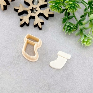 May include: Two plastic Christmas stocking cookie cutters. One is a traditional stocking shape and the other is a more modern, simple stocking shape.
