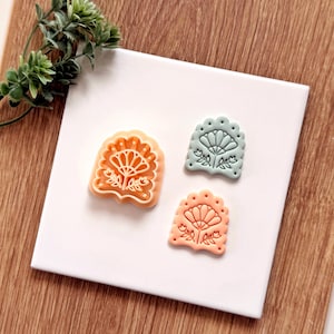 May include: Three decorative cookie cutters in shades of orange and blue-green, each featuring a floral design. The cutters are arranged on a white square, with a sprig of greenery in the background. The cutters are approximately 2 inches in length.