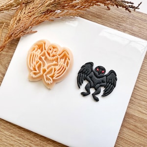 Pode incluir: Duas cortadores de biscoitos com tema de Halloween. Um é um cortador de cor pêssego claro em forma de criatura alada. O outro é uma criatura alada preta com olhos vermelhos. Ambos estão em uma superfície branca.