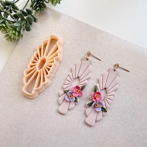Könnte beinhalten: Hellrosa Ohrringe mit goldfarbenen Stiften, die ein fächerförmiges Design aufweisen und mit kleinen rosa und lila Blumen verziert sind. Ein passender Ton-Ausstecher ist ebenfalls vorhanden, was auf ein DIY-Schmuckthema hindeutet.