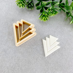 Può includere: Due stampini per biscotti a forma di design geometrico. Lo stampino più grande è fatto di plastica marrone chiaro e ha cinque triangoli sovrapposti. Lo stampino più piccolo è fatto di plastica bianca e ha tre triangoli sovrapposti.